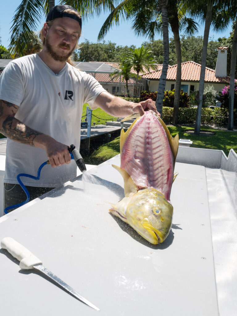 The fishmonger rinses down a filleted yellow jack and the cleaning table.